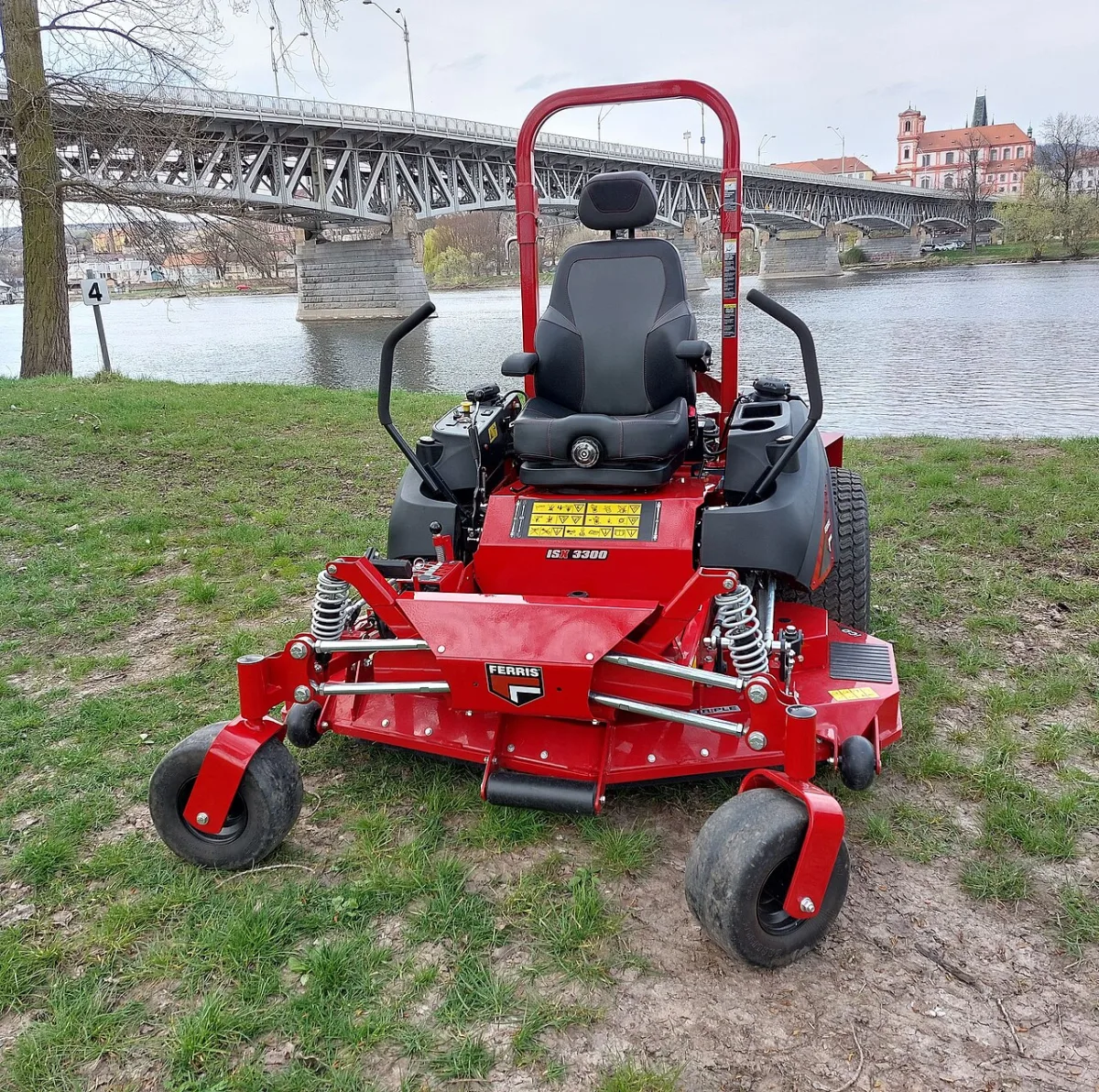 Rode zero-turn zitgrasmaaier met veerophanging op een grasveld bij een rivier met brug op de achtergrond