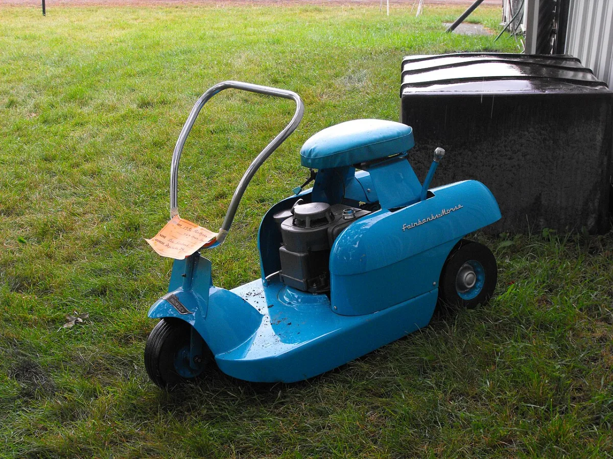 Blauwe kleine zitgrasmaaier met zadel en motor, geparkeerd op gras naast een schuur